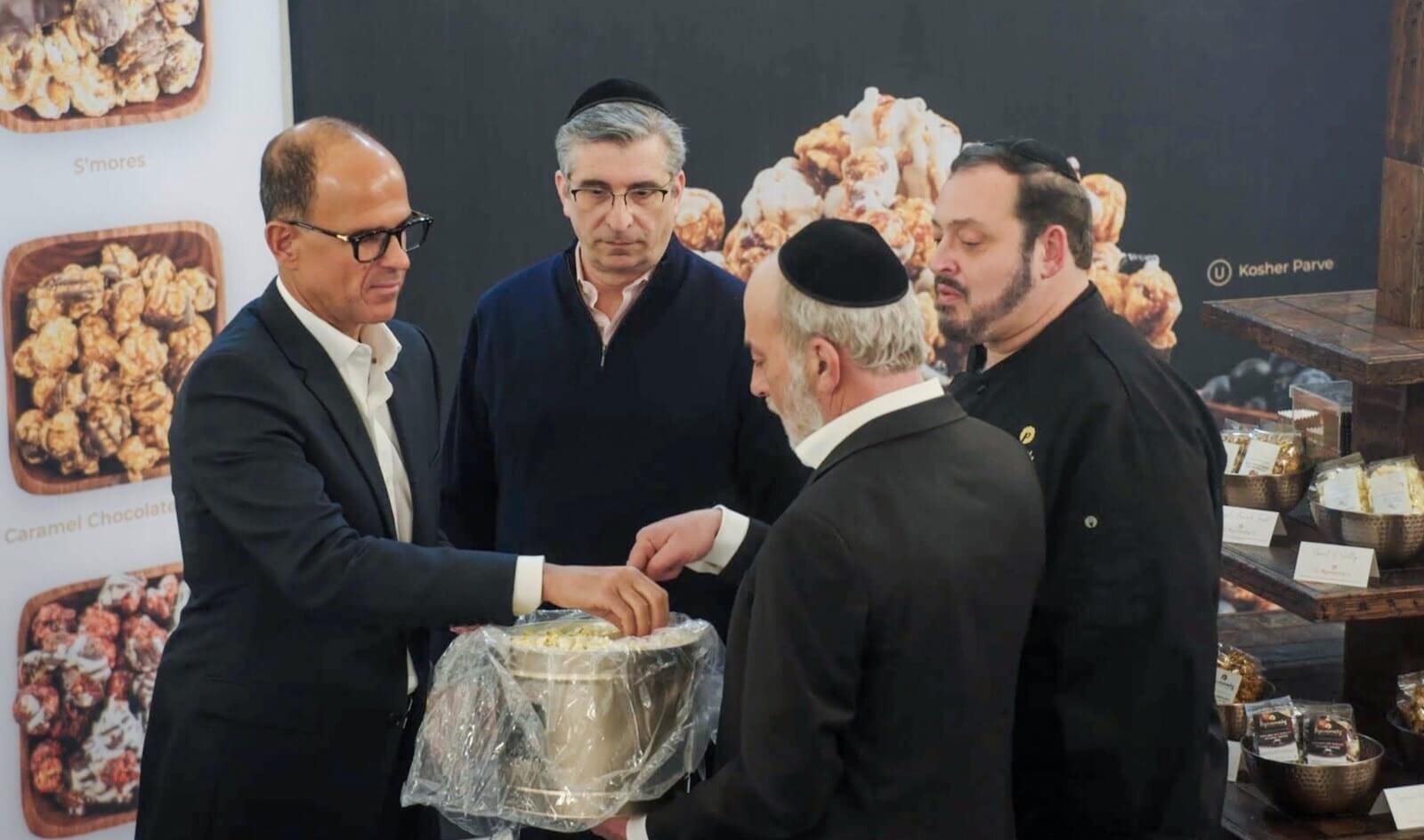 Four men, some wearing kippahs, gather around a bowl of popcorn at a display table featuring various popcorn flavors in a food setting.