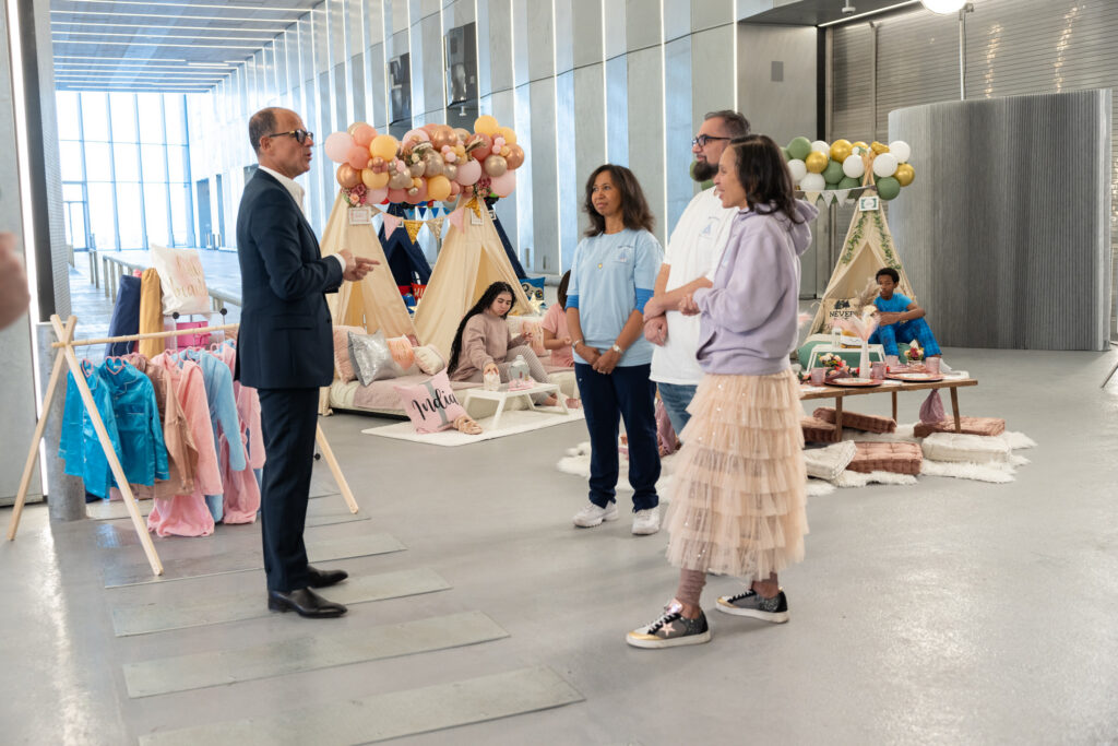 Marcus Lemonis in a suit speaks to three casually dressed adults in front of childrens play tents and decorations inside a modern building.