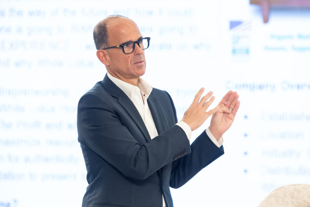 Marcus Lemonis gestures with his hands while giving a presentation in front of a blurred projection screen with text.