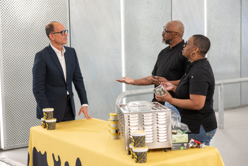 Marcus Lemonis in a suit stands at a counter with two people in black shirts behind it, next to food containers and cups on a yellow tablecloth.