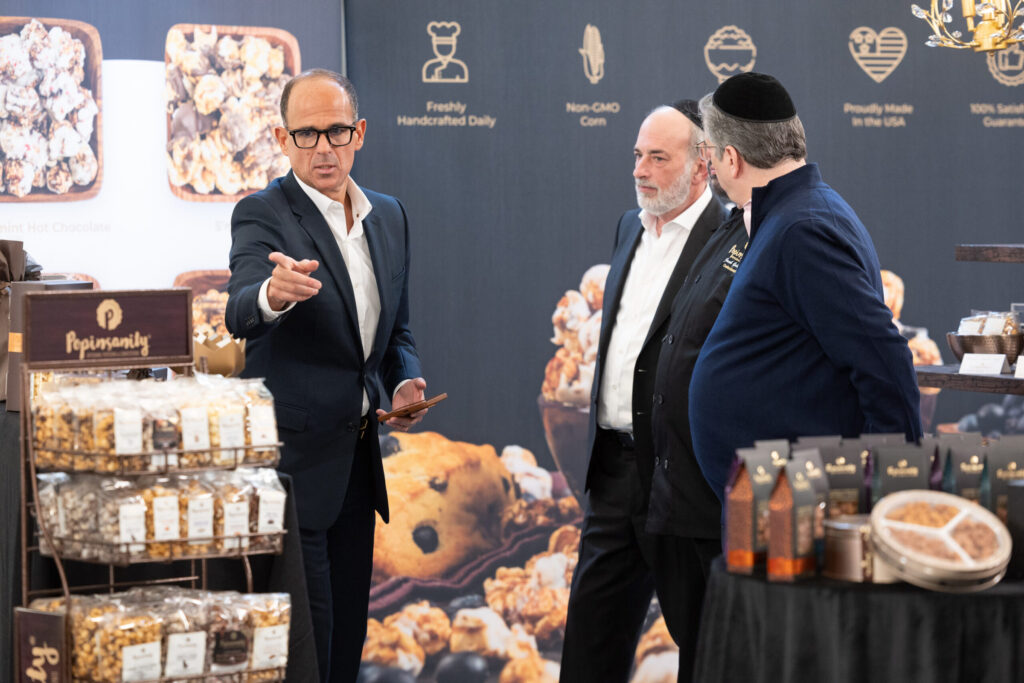 Marcus Lemonis and two men stand and talk near a display of packaged popcorn and snacks at a trade show booth, with branded signage and product displays in the background.
