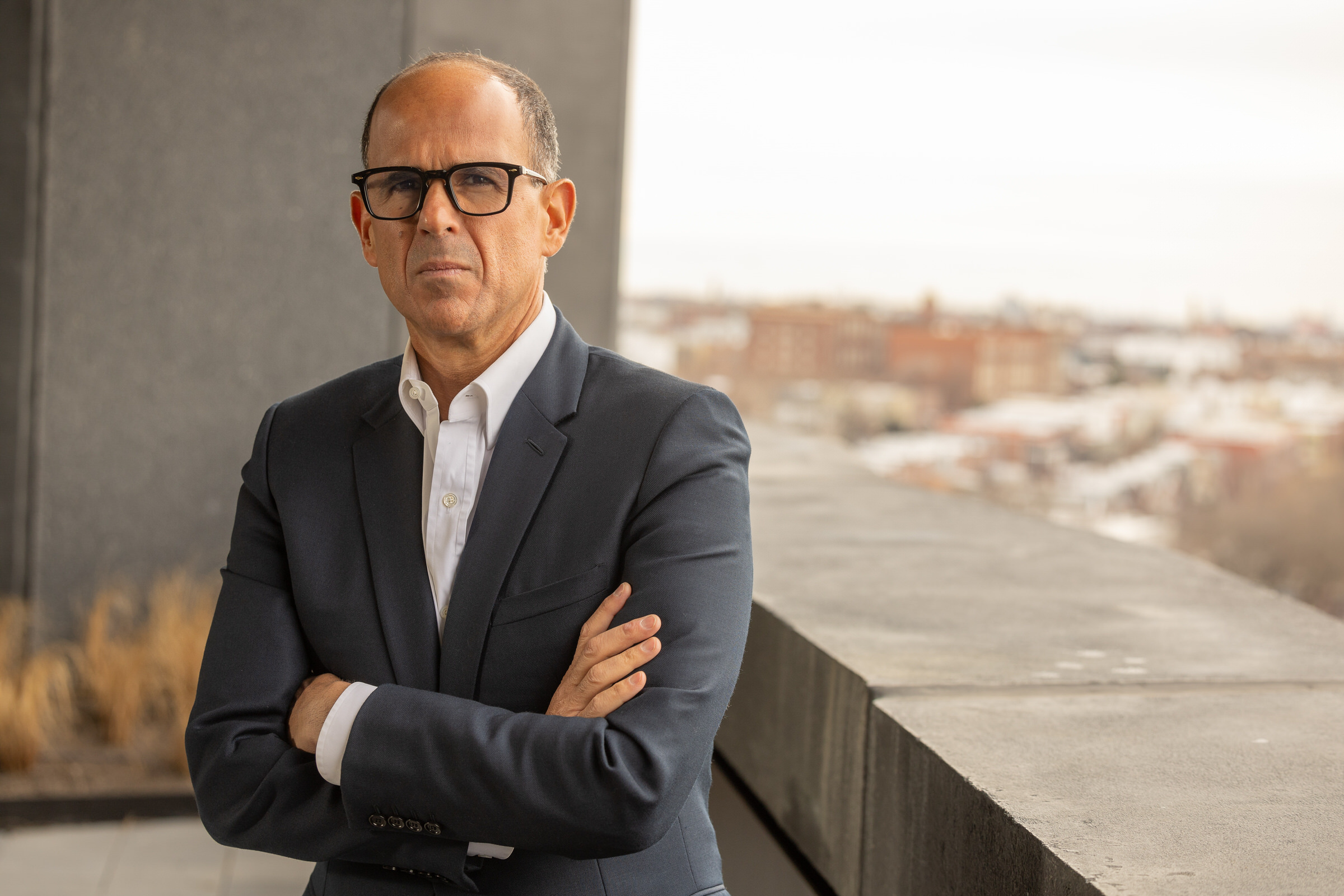 Marcus Lemonis in a dark suit and glasses stands with arms crossed on an outdoor balcony, with a blurred cityscape in the background.