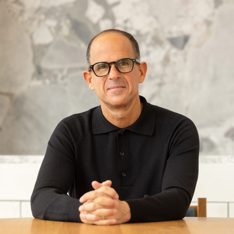 Marcus Lemonis wearing glasses and a black long-sleeve shirt sits at a wooden table with his hands clasped, in front of a light-colored textured wall.