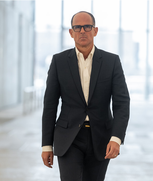 Marcus Lemonis in a dark suit and glasses walks confidently through a bright, modern indoor space.