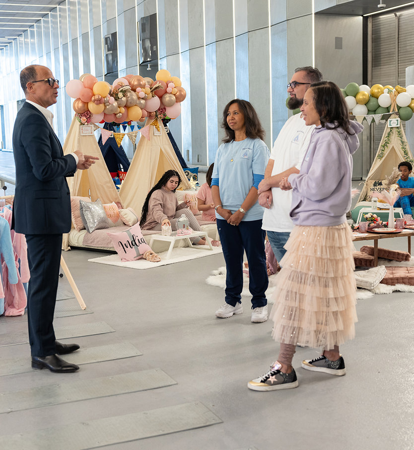Marcus Lemonis talks to three casually dressed adults in front of decorated tents at an indoor event with balloons and picnic setups.