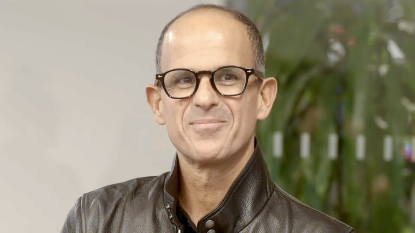 Marcus Lemonis wearing glasses and a black leather jacket is standing indoors with a faint smile; a blurred green plant is visible in the background.