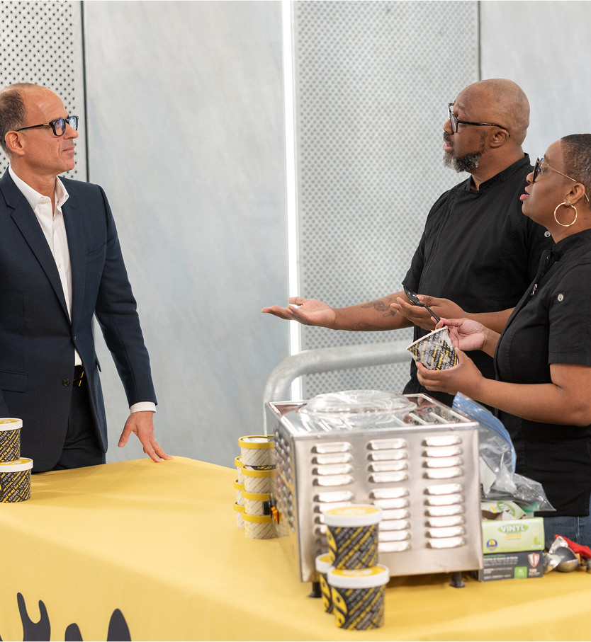 Three people stand at a table with a yellow tablecloth, stacked cups, and a metal food machine, engaged in conversation indoors.