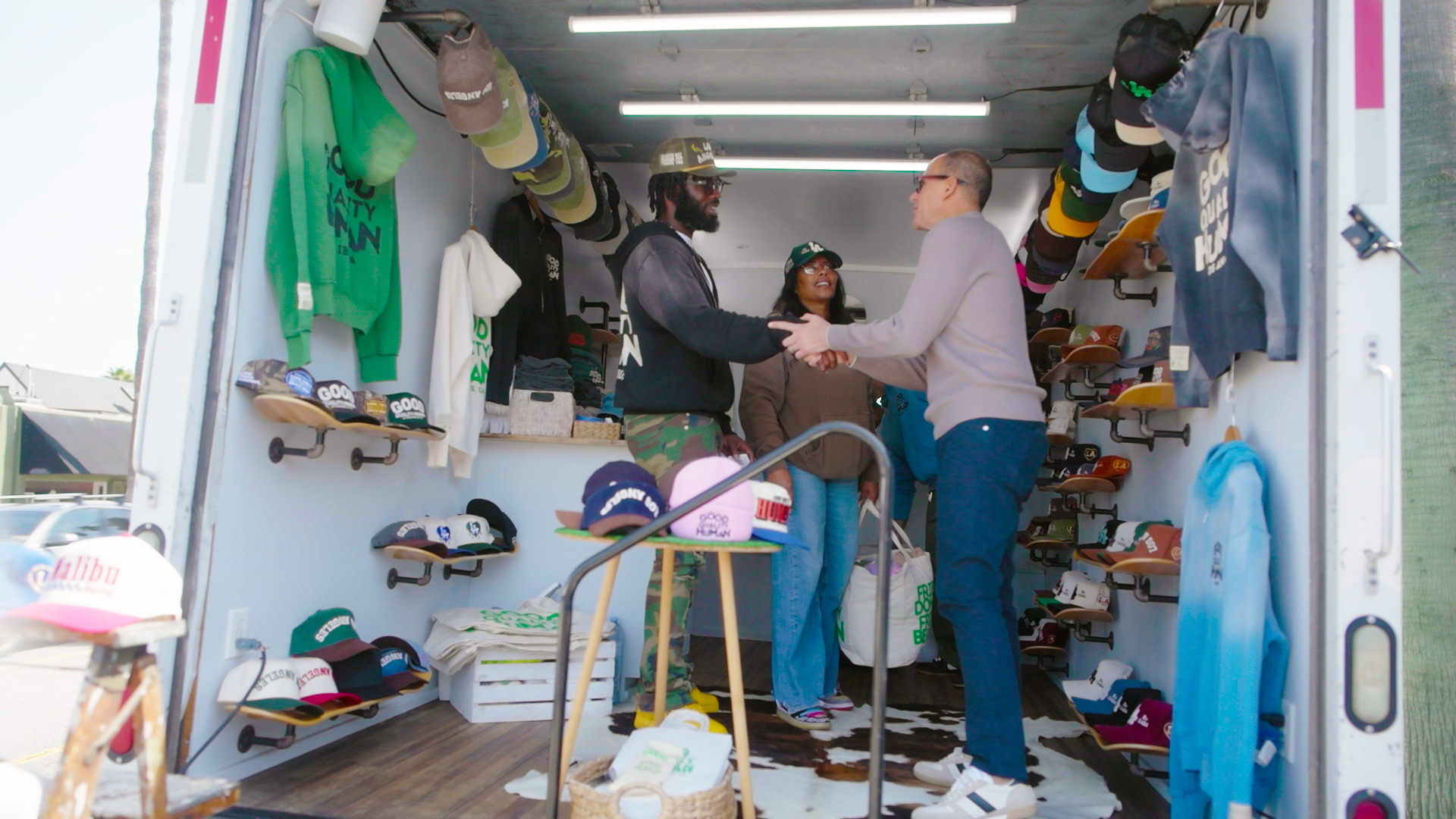Three people stand inside a small shop filled with hats and clothing, with two of them shaking hands near the entrance.