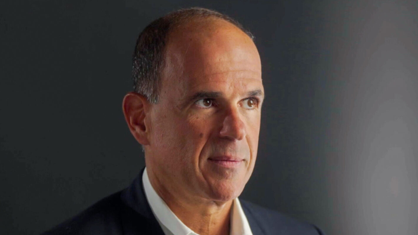Marcus Lemonis wearing a dark suit jacket and white shirt, standing against a dark background.