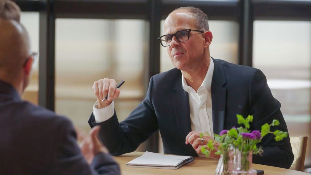 Marcus Lemonis in a suit and glasses sits at a table with a notebook and pen, listening to another person across from him. A small vase with flowers is on the table.