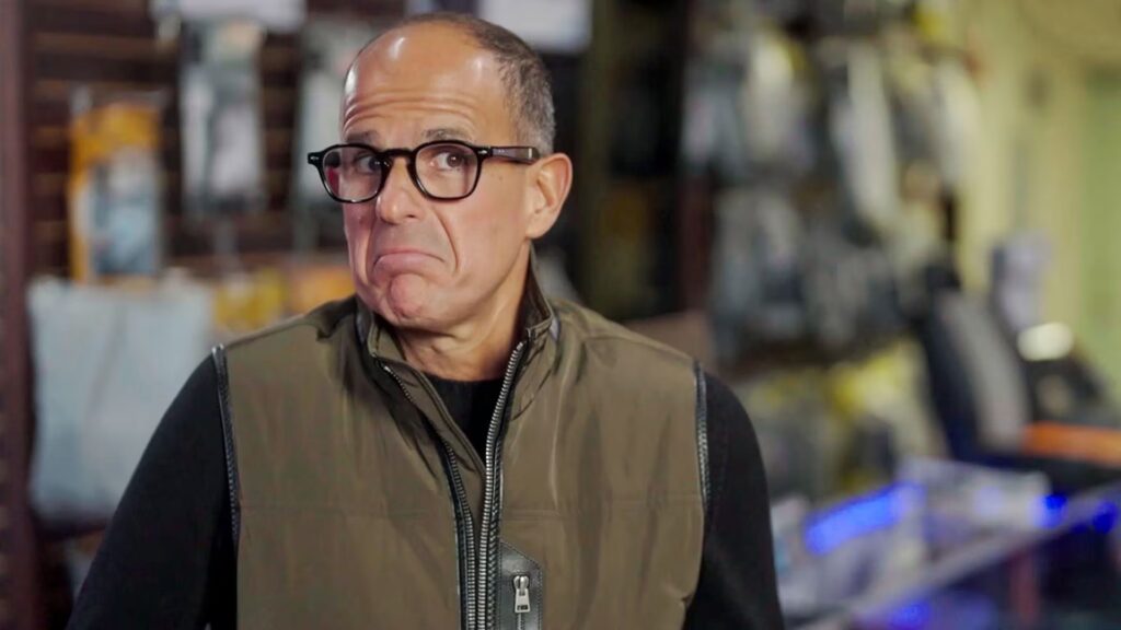 Marcus Lemonis glasses and a vest stands indoors, looking to the side with a skeptical or uncertain expression.