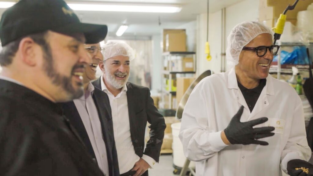 Four men, three in business attire and one in a lab coat and hairnet, stand together smiling and conversing in an industrial or laboratory setting.