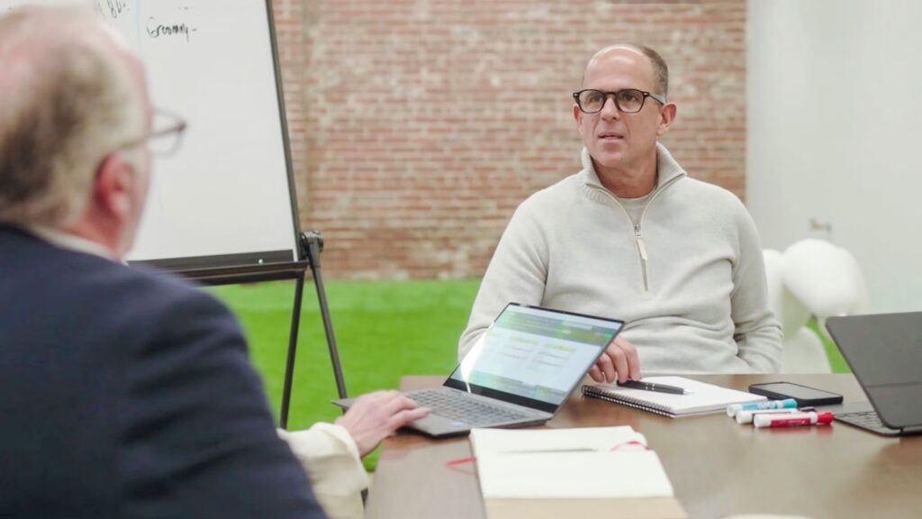 Two men sit at a table with laptops, notebooks, and markers, having a discussion in a modern office with a brick wall and whiteboard visible.