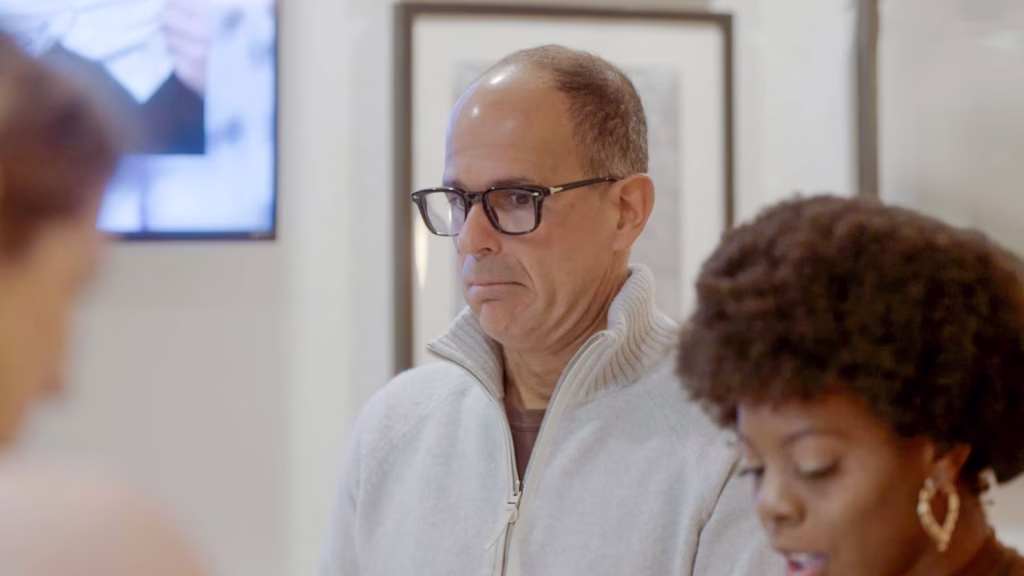 Marcus Lemonis wearing glasses and a light gray zip-up sweater stands indoors, looking ahead, with two people partially visible in the foreground.