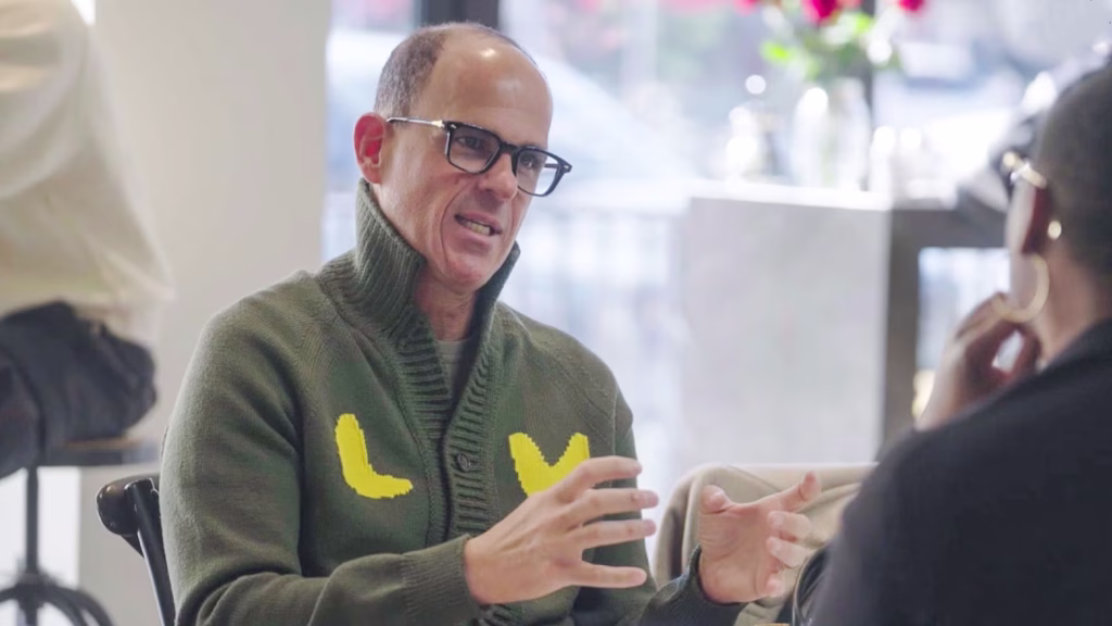 Marcus Lemonis wearing glasses and a green sweater with yellow shapes gestures while talking to another person in a well-lit indoor setting.