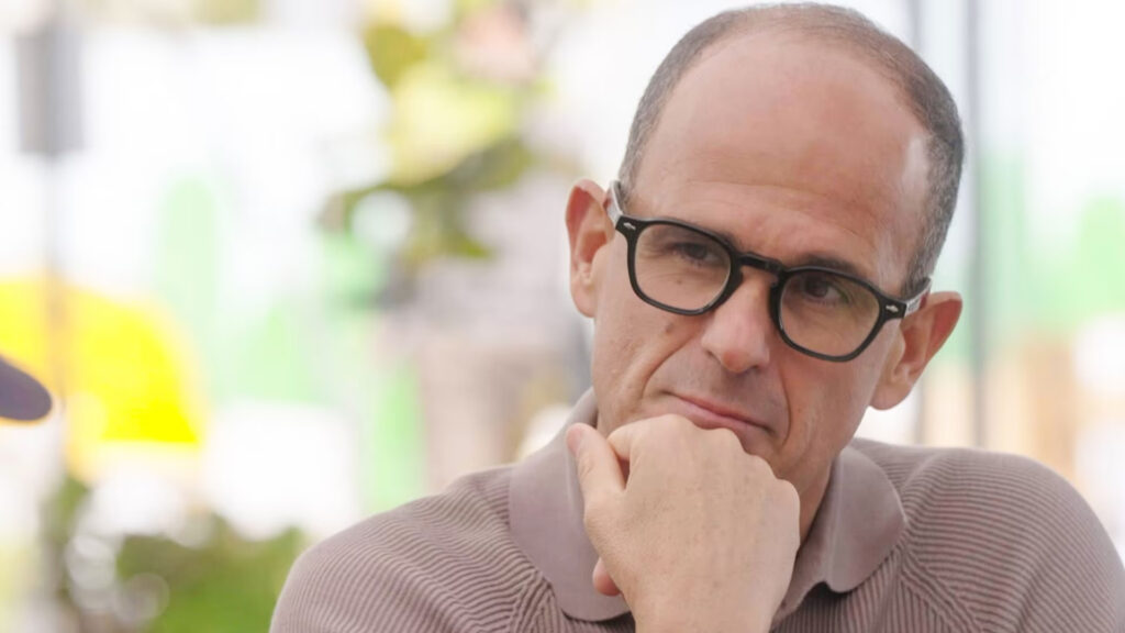 Marcus Lemonis rests his chin on his hand, looking thoughtful. He is wearing a light brown collared shirt.